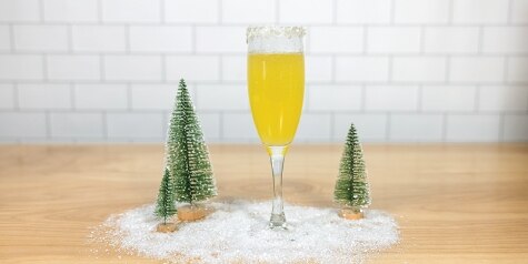 A champagne flute surrounded by Christmas decoration holds a light orange Snowbirds cocktail, garnished with a coconut rim.