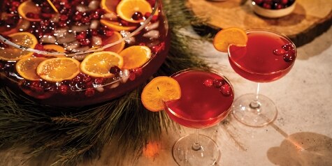 A glass punch bowl holds red Berry & Bright Punch, garnished with orange slices and cranberries - two coupe glasses in front.