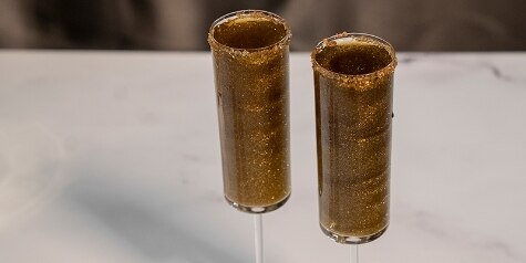 Two flute glasses hold golden Meet Me at Midnight cocktails, garnished with edible glitter and gold sanding sugar.