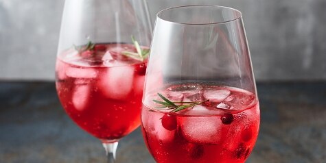 Two wine glasses on an industrial surface hold red Cranberry Rosemary Spritz cocktails, garnished with rosemary and cranberries.
