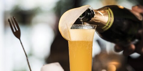 A champagne flute holds an orange-colored Pear Mimosa being topped with sparkling wine, garnished with a pear slice.