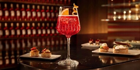 A deep orange Campari Spritz sits on a table with plates of desserts.