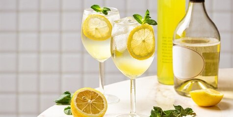 Two yellow Limoncello Spritzes on a white marble tabletop, with lemons, mint sprigs and bottles of prosecco and limoncello.