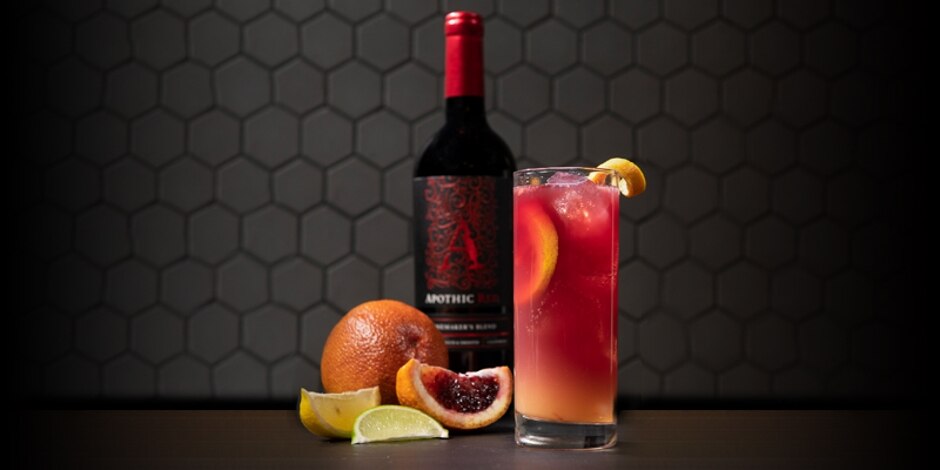 Orange cocktail in ice-filled highball glass, garnished with lemon twist in front of a bottle of Apothic wine and orange wedges.