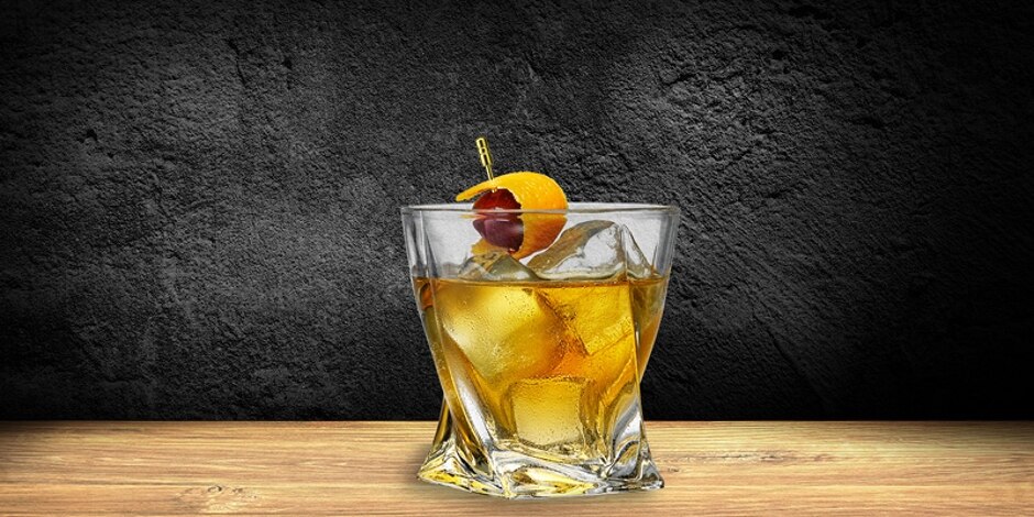 A tequila old fashioned in a rocks glass with ice, garnished with skewered orange peel and cherry, sits on a wooden table.