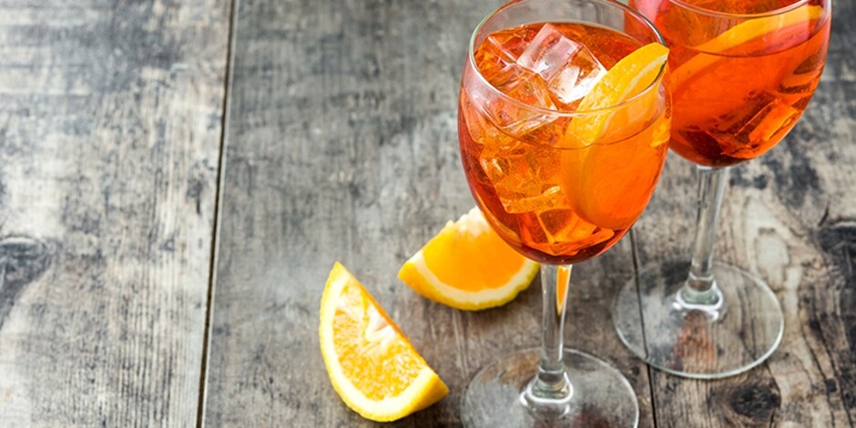 Two wine glasses filled with the vibrant orange Aperol Spritz garnished with orange slices, sit on a gray washed table.