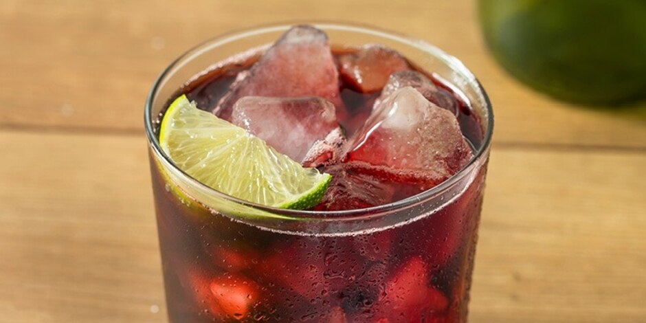 Blueberry Rebel Spritzer, dark red cocktail, on the rocks in a rocks glass, garnished with a lime wedge.