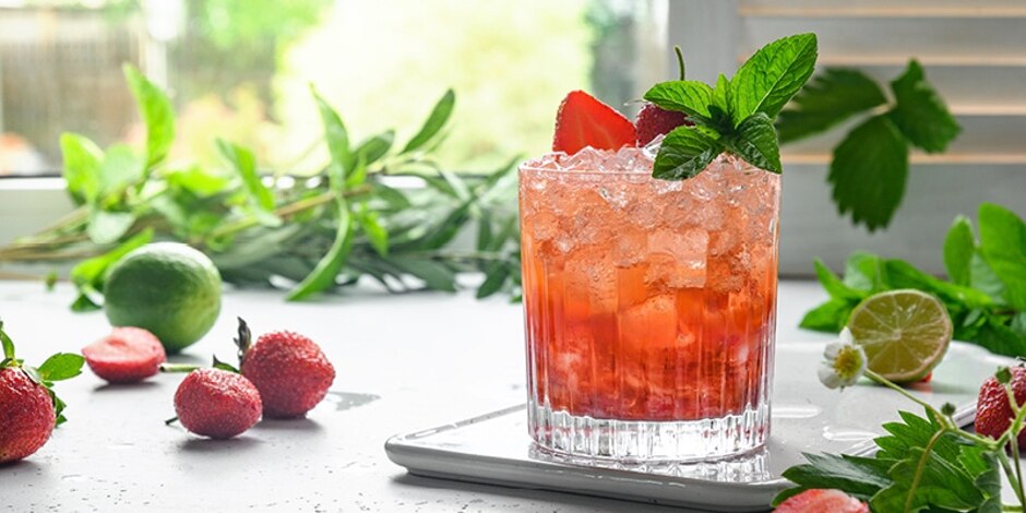 Red cocktail with strawberry and mint garnish on a table with strawberries, limes and mint.