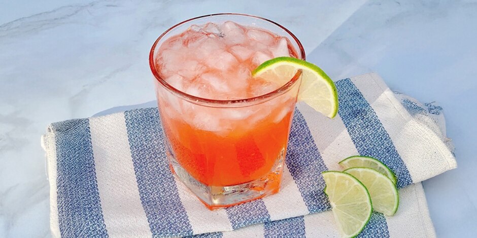 An orange-colored Italian Margarita Spritz on a blue-and-white striped tablecloth, accompanied by lime slices.