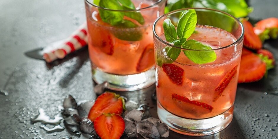 Two light pink cocktails with strawberry slices and basil leaves in rocks glasses, sitting on a black tabletop.