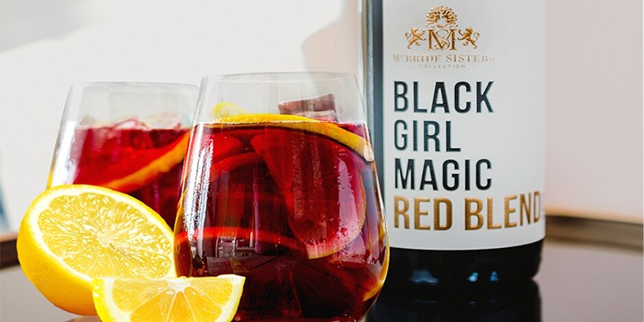 Red cocktail in wine glass filled with ice and lemon slices, next to a bottle of Black Girl Magic red wine.