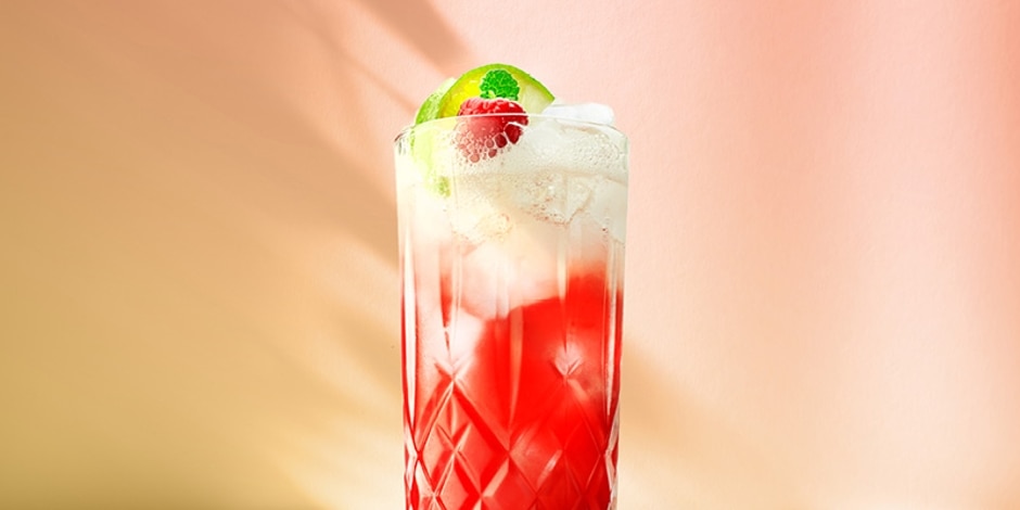 The Floradora cocktail, served in a tall glass, has a raspberry hue topped with ginger beer, garnished with raspberry and lime.