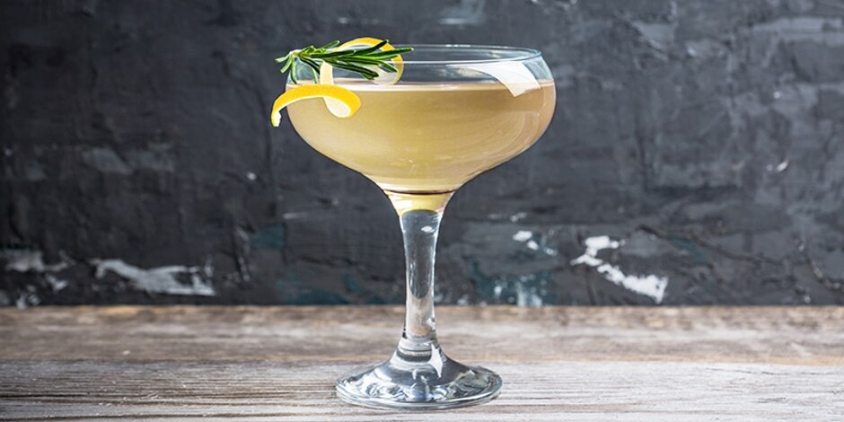 A coupe glass filled with a pale yellow-brown Smoke & Bubble cocktail garnished with a lemon twist and rosemary sprig.