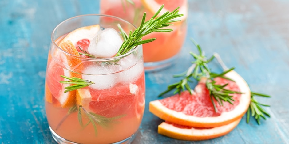 Virgin Paloma in a glass with grapefruit wedges, rosemary sprig and ice on a blue wood table next to grapefruit and rosemary.