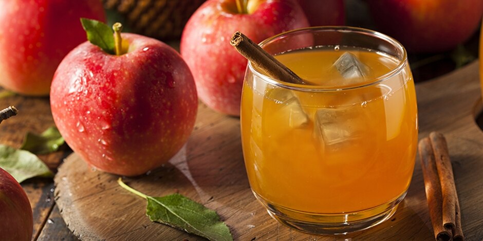 Apple Cider Bourbon cocktail garnished with a cinnamon stick, sitting on a wooden table with apples in the background.