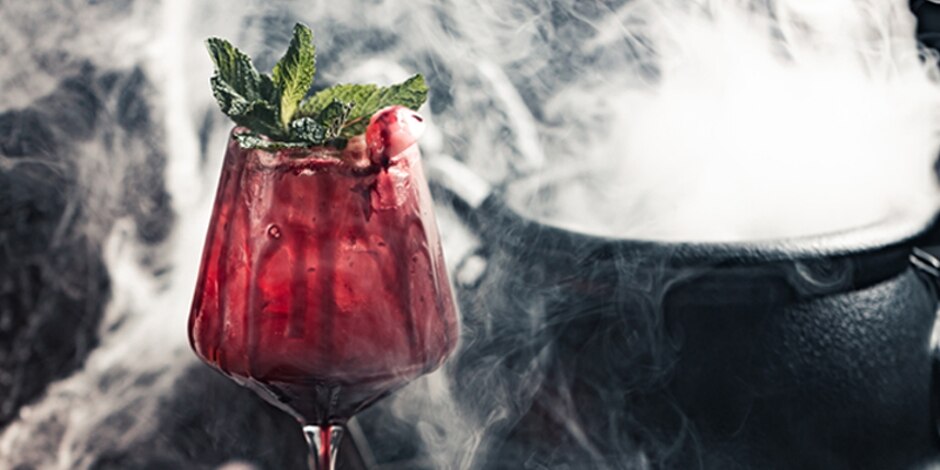 A wine glass holds a blood red Dracula’s Kiss cocktail, garnished with a mint sprig, surrounded by smoke.